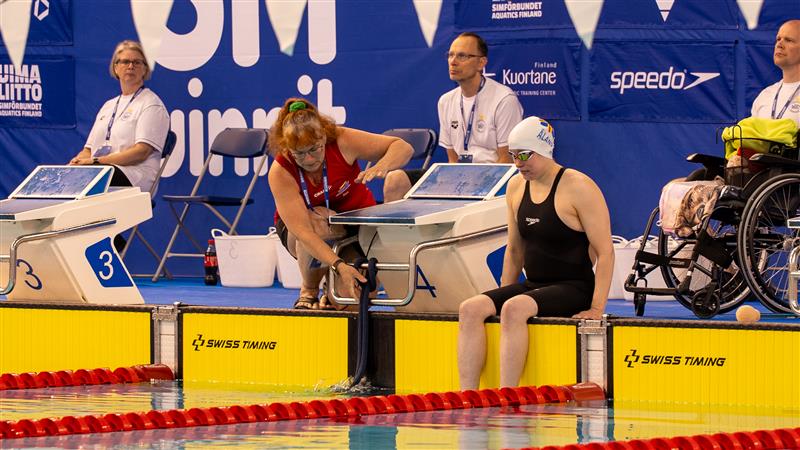 Liikuntavammainen uimari istumassa uimaradan päädyssä lähtötelineen vieressä. Taustalla pyörätuoli, josta on noustu pois.